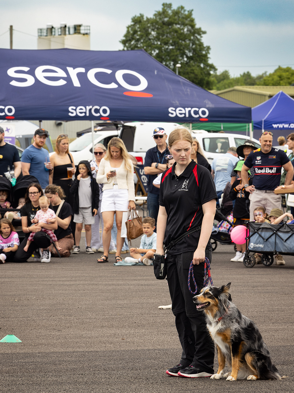 A female trainer standing next to a sitting dog on the tarmac during a public demonstration, with a crowd of spectators and a large blue "Serco" event tent in the background.